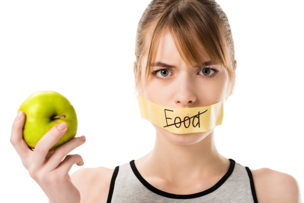 mujer joven con cinta adhesiva con alimentos tachados a través de la palabra que cubre la boca sosteniendo manzana aislada en blanco
 - Foto, Imagen