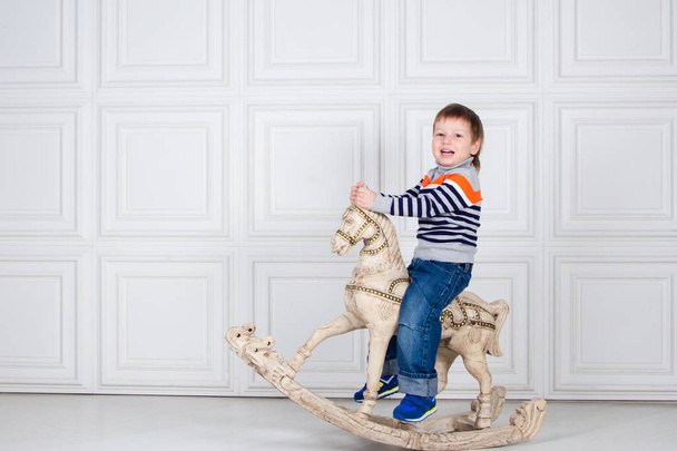 little boy swinging on wooden horse. funny three-year-old boy in jeans and sweater on white background. Carefree childhood - Φωτογραφία, εικόνα