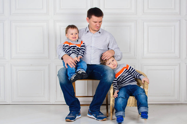 dad with two children on his knees sitting on the chair in front of white wall. Lifestyle. Children in identical sweaters - Φωτογραφία, εικόνα