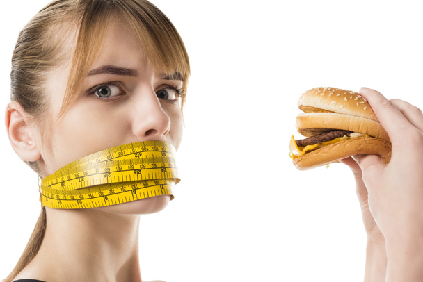 young woman with mouth tied in measuring tape trying to eat burger isolated on white - Zdjęcie, obraz