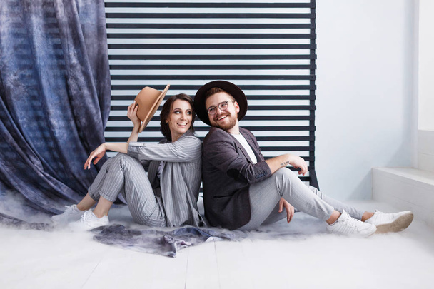 座っている陽気な若いカップルは、幸せそうに笑って床に互いにバックアップします。背景の黒と白のストライプ. - 写真・画像