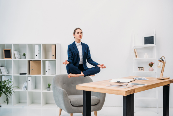young businesswoman meditating with closed eyes while levitating at workplace  - Foto, afbeelding
