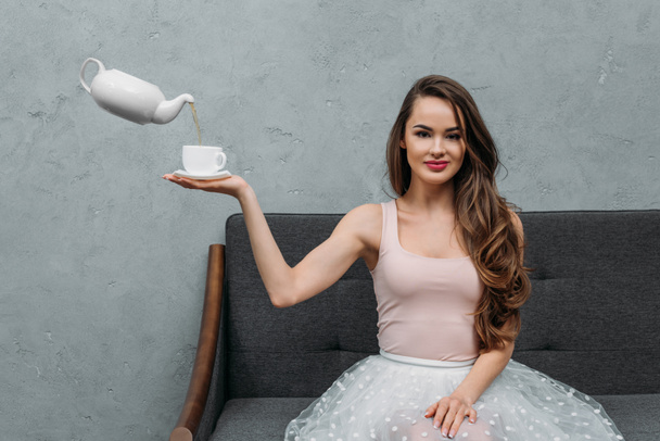 hermosa mujer sonriendo a la cámara y sosteniendo la taza mientras levita tetera vertiendo té
 - Foto, imagen