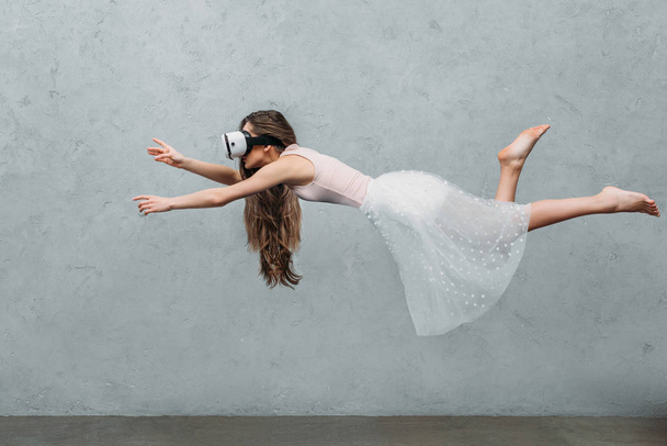 young barefoot woman in virtual reality headset levitating on grey  - 写真・画像
