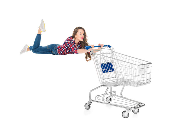 Lächelnde junge Frau schwebt mit leerem Einkaufswagen auf weißem Grund - Foto, Bild