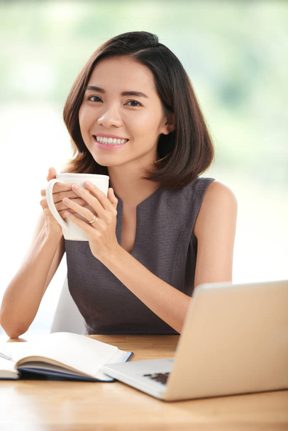 hermosa estudiante asiática chica sosteniendo taza de té y mirando a la cámara mientras está sentado en la mesa con libro de texto y portátil
 - Foto, Imagen