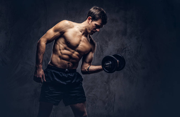 Retrato de estudio de un hombre sin camisa atlético tatuado vestido con pantalones cortos deportivos, haciendo ejercicios sobre bíceps sobre fondo oscuro
. - Foto, imagen