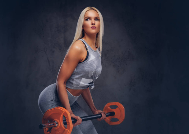 Спортивная блондинка фитнес-женщина делает упражнения с штангой на сером фоне
. - Фото, изображение