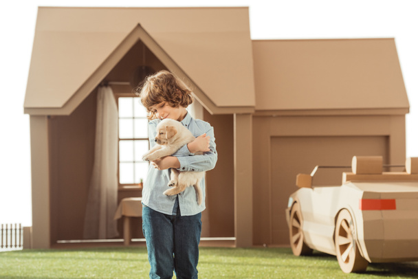 little kid holding his adorable labrador puppy in front of cardboard house isolated on white - Photo, Image