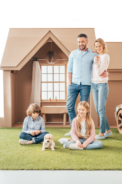 young family with cute puppy on yard of cardboard house isolated on white - Photo, Image