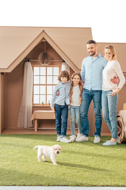 happy young family with adorable puppy on yard of cardboard house isolated on white - Φωτογραφία, εικόνα