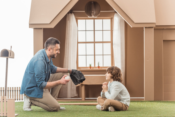 father teaching his son how to play baseball on grass in front of cardboard house - Photo, Image