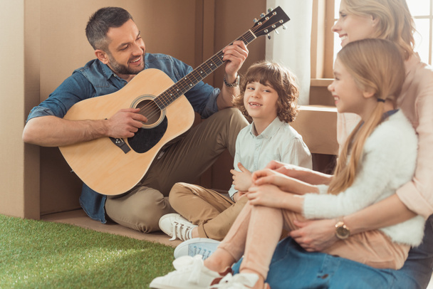 счастливый красивый отец играет на гитаре для детей и жены в новом картонном доме
 - Фото, изображение