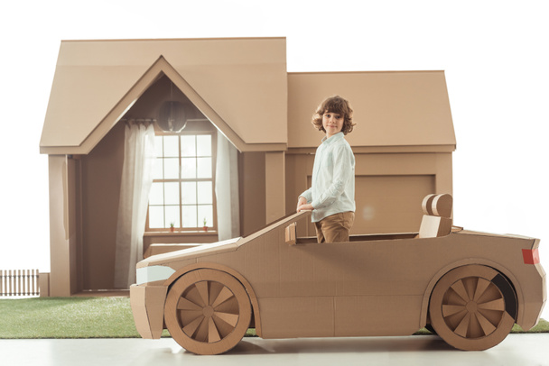 μικρό παιδί στέκεται στο αυτοκίνητο από χαρτόνι μπροστά από χαρτόνι σπίτι που απομονώνονται σε λευκό - Φωτογραφία, εικόνα