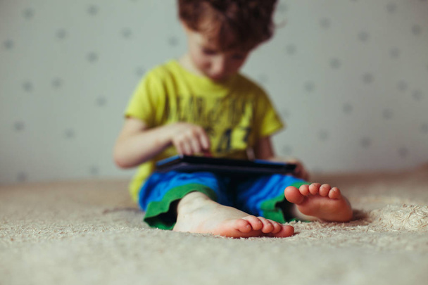 Kid with tablet sitting on bed and watching cartoons. Selective Focus on the feet. Cute boy in green t-shirt and blue pants - Photo, image