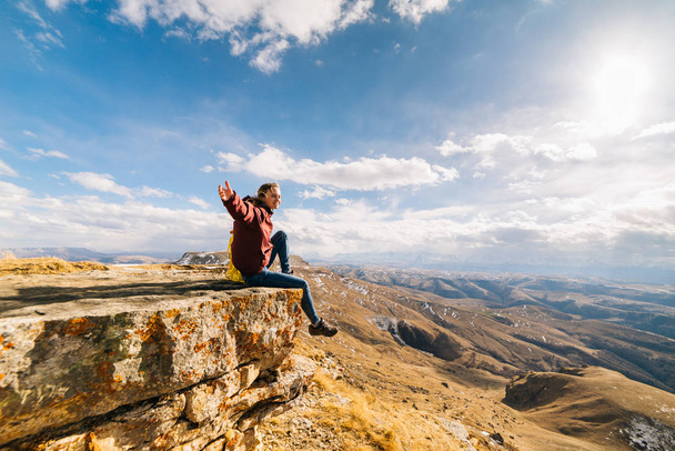 τουριστική ευτυχισμένη γυναίκα κάθεται πάνω σε ένα βράχο στο παρασκήνιο των βουνών σε μια ηλιόλουστη ημέρα - Φωτογραφία, εικόνα