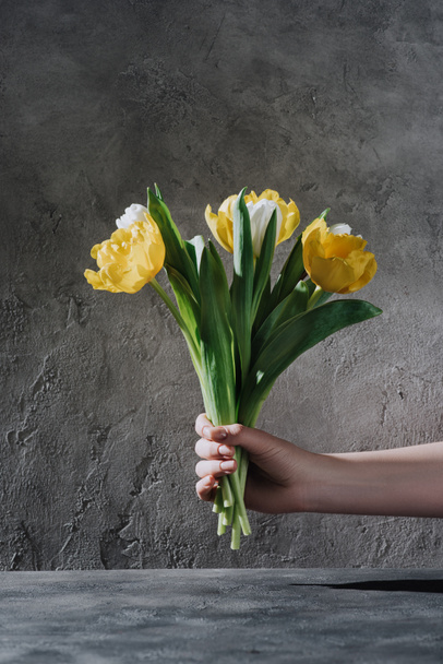 abgeschnittene Ansicht einer Frau mit gelben und weißen Tulpenblüten auf grauer Oberfläche - Foto, Bild