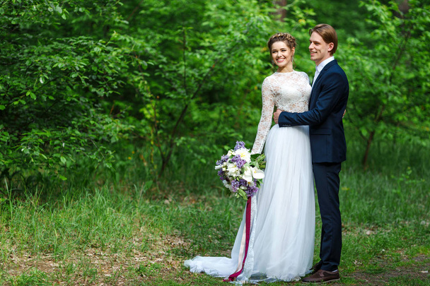 Young beautiful groom in dark blue suit and bride in white crop top dress and holding big wedding bouquet with lilac and long ribbons. Happy couple on spring wedding walk - Фото, зображення