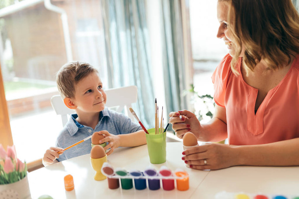 Happy young mother and son are sitting at a table painting Easter eggs - Photo, image