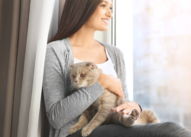 Jonge vrouw met schattig huisdier kat, zittend op de vensterbank thuis - Foto, afbeelding