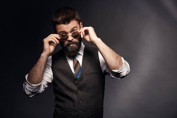 Studio portrait of attractive handsome man with dark hair, mustache and beard in white shirt, brown vest and colorful tie takes off round sunglasses - 写真・画像
