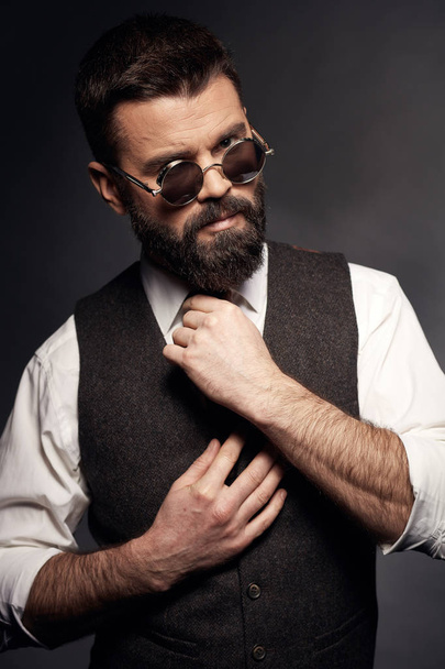 Studio portrait of attractive handsome man with dark hair, mustache and beard in white shirt, brown vest, colorful tie and round sunglasses - Foto, imagen