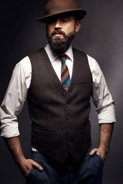 Studio portrait of attractive handsome man with dark hair, mustache and beard in white shirt, brown vest, colorful tie and brown hat. Hands in pockets - Foto, Bild