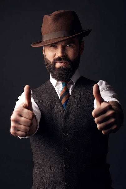 Studio portrait of handsome man with dark hair and mustache in white shirt, brown vest, colorful tie and brown hat with thumbs up. Old fashioned style, gentleman. - Foto, Imagem