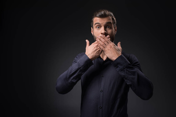 portrait of shocked man covering mouth with hands isolated on black - Fotografie, Obrázek