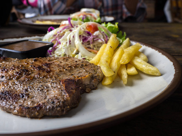close up of T-bone steak with salt and pepper, golden french fries and green vegetables. on white plate, Fork and knife, wooden table in the restaurant - Photo, Image