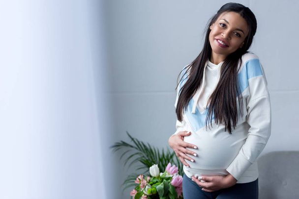 窓に立っている彼女の腹を見て幸せなアフリカ系アメリカ人の妊娠中の女性 - 写真・画像