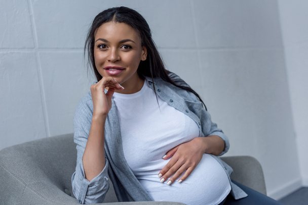улыбающаяся африканская беременная женщина смотрит в камеру, сидя на кресле
 - Фото, изображение