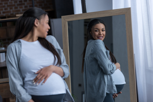 african american pregnant woman looking at her reflection in mirror - Photo, Image