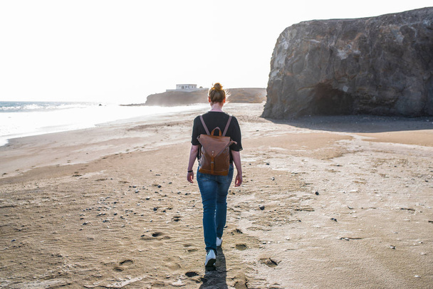 fiatal szőke nő alkalmi ruhákat és bőr hátizsák séta kihalt szeles tengerparton - Fotó, kép