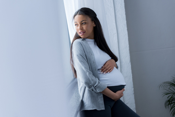 beautiful african american pregnant woman touching her belly while looking at window - Foto, immagini