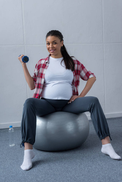 african american pregnant woman sitting on fit ball and training with dumbbell - Fotografie, Obrázek