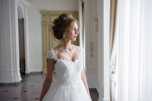 όμορφη νύφη σε ένα νυφικό σε ένα πολυτελές λευκό δωμάτιο με έναν μεγάλο πολυέλαιο - Φωτογραφία, εικόνα