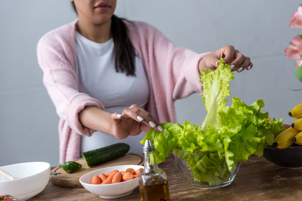 vista cortada da mulher grávida afro-americana fazendo salada fresca na cozinha
 - Foto, Imagem