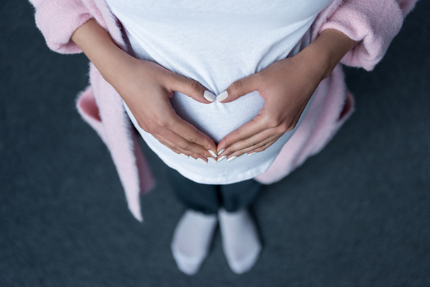 обрізаний вид вагітної дівчини, що робить символ серця руками на животі
 - Фото, зображення