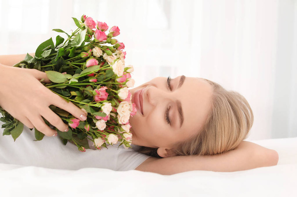 Beautiful young woman with bouquet of roses on bed at home - 写真・画像