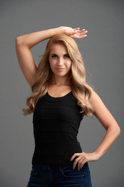 Porträt eines selbstbewussten blonden Models mit schwarzem Top, das vor grauem Hintergrund posiert - Foto, Bild