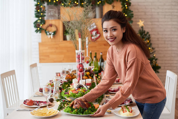 Beautiful Vietnamese young woman putting dish on Christmas table and smiling at camera - Foto, Bild
