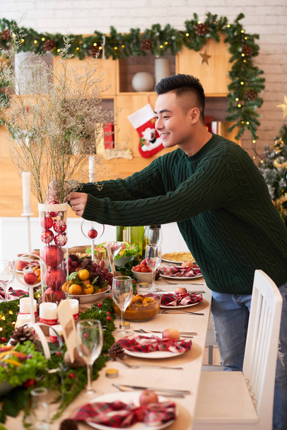 Vietnamese young man putting vase with baubles on Christmas dinner table - Photo, Image