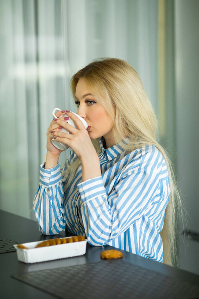 beautiful young blonde girl at home in the kitchen drinking coffee at breakfast - Fotó, kép