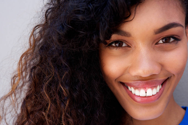 Nahaufnahme Porträt der schönen jungen afrikanisch-amerikanischen Frau mit lockigem Haar, die vor grauem Hintergrund lächelt - Foto, Bild