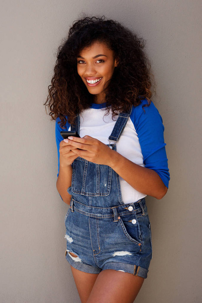 Retrato de una hermosa joven afroamericana con cabello rizado sosteniendo un teléfono inteligente contra un fondo gris
 - Foto, imagen