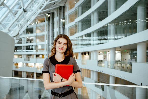 The young business woman in gray office dress holds in hand tablet with documents. The girl costs against the background of office premises of business center. - Foto, immagini