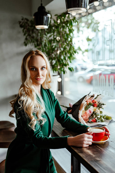 die schöne junge Frau im Kaffeehaus bei einem Termin. Die blauäugige Blondine im grünen Anzug sitzt an einem kleinen Tisch im Café und hält eine Tasse Kaffee in der Hand. in der Nähe auf einem Tisch ein Strauß Blumen. - Foto, Bild