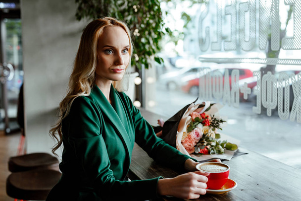 The beautiful young woman in coffee house on an appointment. The blue-eyed blonde in a green suit sits at a little table in cafe and holds a cup of coffee in hand. Nearby on a table a bouquet of flowers. - Фото, изображение