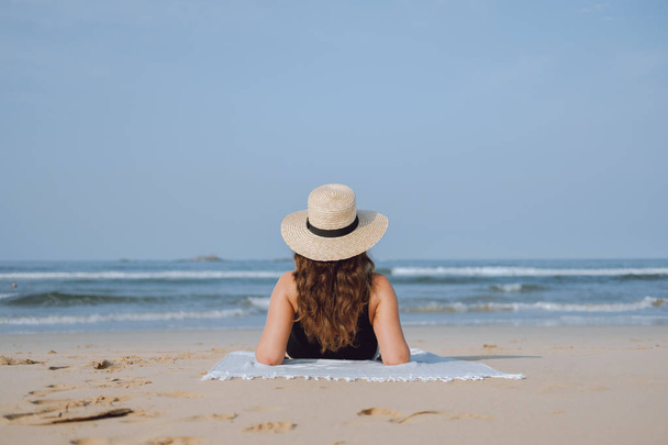 Back view. Girl in a straw hat looks at the ocean sunbathing on the sand - Fotografie, Obrázek